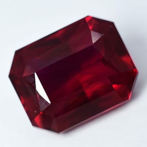 May include: A rectangular-cut ruby gemstone with multiple facets. The gem is a deep red colour with internal light reflections. The cut creates a brilliant appearance. The gemstone is set against a white background.