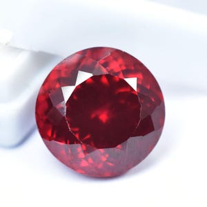 Könnte beinhalten: Ein facettierter, rund geschliffener, tiefroter Edelstein. Der Edelstein hat eine brillante, reflektierende Oberfläche mit mehreren Facetten, die das Licht einfangen. Der Stein ist vor einem weißen Hintergrund platziert, was seine satte Farbe und Klarheit hervorhebt. Dieser Edelstein ist für Schmuck geeignet.