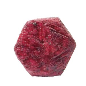 Pode incluir: Uma pedra preciosa de rubi bruta e não cortada. O rubi é de uma cor vermelha profunda e rica com uma superfície áspera e texturizada. A pedra preciosa tem uma forma hexagonal e está sobre um fundo branco.