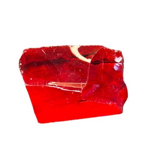 May include: A vibrant, irregularly shaped piece of red glass. The glass has a glossy, reflective surface with a few white markings. The edges are rough and uneven, suggesting it may be a broken piece or a natural formation.