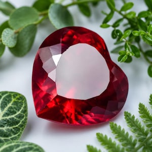 May include: A heart-shaped, faceted gemstone in a deep red colour. The gem is surrounded by green foliage, creating a striking contrast. The gem's cut reflects light, highlighting its shape and colour. A beautiful piece for jewellery or a collection.
