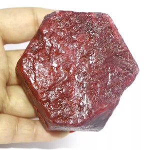 Op de afbeelding: Een grote, dieprode robijn edelsteen met een gefacetteerde, zeshoekige vorm. De robijn heeft een glad, gepolijst oppervlak en is een rijke, levendige rode kleur.