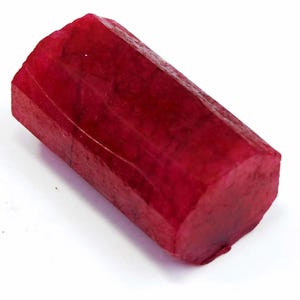 May include: A raw, uncut ruby gemstone with a deep red color. The rectangular stone has a faceted shape and a slightly rough texture. The ruby is displayed against a white background, highlighting its rich color and natural form.