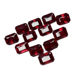 May include: A collection of twelve red gemstone cabochons, cut in a rectangular emerald shape. The gemstones are arranged in a triangular pattern on a white background.