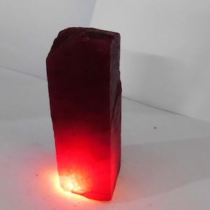 Puede incluir: Una gran gema de rubí en bruto, de color rojo intenso. El rubí rectangular tiene una superficie rugosa y texturizada. La parte inferior de la gema brilla con una luz roja brillante, creando un efecto degradado.