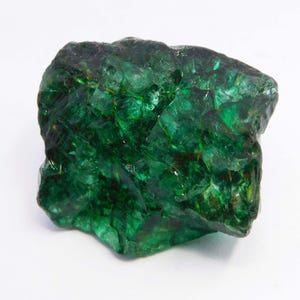 Può includere: Una gemma grezza verde smeraldo con una struttura cristallina. La superficie riflette la luce, mostrando varie sfumature di verde e un aspetto strutturato. La gemma è posta su uno sfondo bianco.