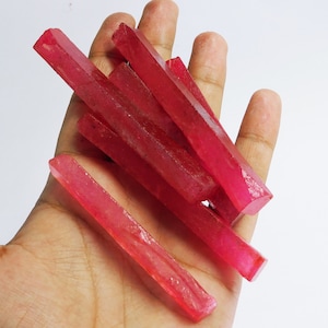 May include: A collection of eight raw ruby crystals, each with a long, rectangular shape and a deep red color. The crystals are arranged in a pile, with some overlapping.
