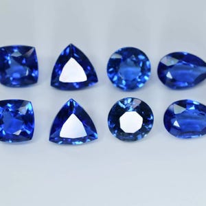 Könnte beinhalten: Acht blaue Edelsteine in verschiedenen Formen und Größen, darunter runde, birnenförmige und dreieckige Schliffe. Die Edelsteine sind in zwei Reihen zu je vier auf einer weißen Oberfläche angeordnet.