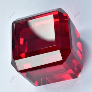 May include: A deep red gemstone cut into a square shape with bevelled edges. The gemstone is faceted and reflects light.