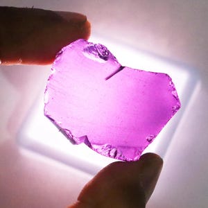Puede incluir: Una gema transl&uacute;cida, de forma irregular, de color p&uacute;rpura vibrante. La piedra se sostiene entre dos dedos, mostrando sus bordes irregulares y su claridad interna. La gema est&aacute; iluminada desde abajo, resaltando su color y textura.