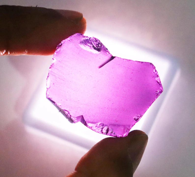 Puede incluir: Una gema transl&uacute;cida, de forma irregular, de color p&uacute;rpura vibrante. La piedra se sostiene entre dos dedos, mostrando sus bordes irregulares y su claridad interna. La gema est&aacute; iluminada desde abajo, resaltando su color y textura.