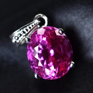 May include: A faceted, oval-cut, pink gemstone pendant with a silver-colored bail. The gemstone is a vibrant magenta color, and the bail is adorned with small, round embellishments. The pendant is set against a dark background.