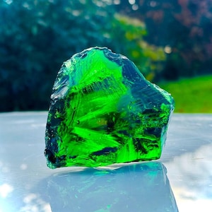 Puede incluir: Un gran fragmento de vidrio verde, con una forma rugosa e irregular. El fragmento refleja la luz, creando un efecto brillante.