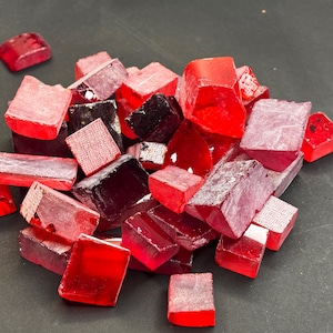 May include: A collection of small, irregularly shaped red glass tiles. The tiles vary in shade from deep crimson to light pink, with some appearing translucent. The pieces are scattered on a dark surface, creating a vibrant display of color and texture.