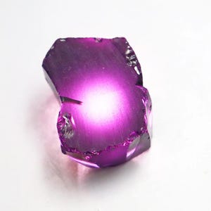 Puede incluir: Una gema de color p&uacute;rpura oscuro, facetada e irregular, con un centro brillante y luminoso. Los bordes de la piedra son &aacute;speros y texturizados, y el aspecto general es transl&uacute;cido. La gema se encuentra sobre un fondo blanco.