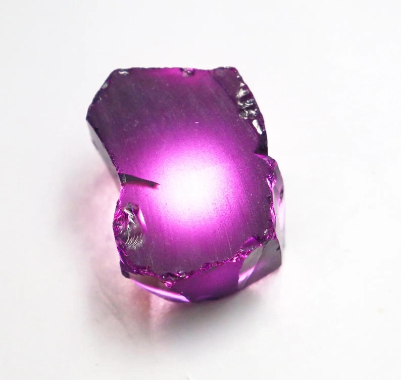 Puede incluir: Una gema de color p&uacute;rpura oscuro, facetada e irregular, con un centro brillante y luminoso. Los bordes de la piedra son &aacute;speros y texturizados, y el aspecto general es transl&uacute;cido. La gema se encuentra sobre un fondo blanco.