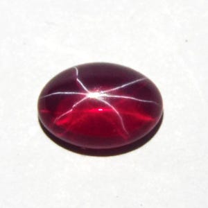May include: A deep red, oval-shaped cabochon gemstone featuring a distinct six-rayed star effect known as asterism. The surface is smooth and polished, displaying a vibrant crimson hue with white light reflecting in a star pattern across the center.