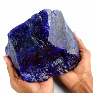 May include: A large, rough, dark blue sapphire gemstone. The sapphire is a deep, rich blue color with purple hues. It is a natural gemstone and has a rough, unpolished surface.
