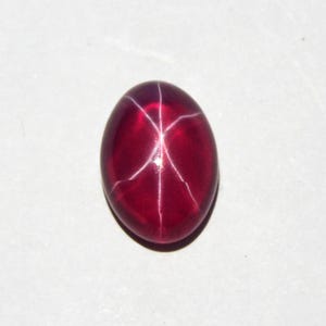 May include: A polished, oval-shaped, deep red gemstone with a six-rayed star pattern. The star effect is created by intersecting white lines across the surface of the stone. The gem is set against a white background.
