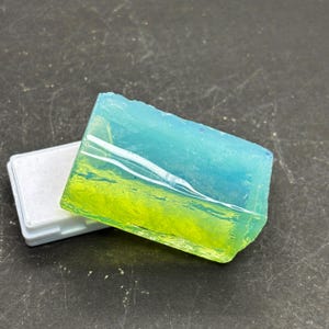 May include: A translucent blue and green soap bar with a yellow layer in the middle. The soap is in a white plastic container.