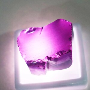 Puede incluir: Una vibrante gema p&uacute;rpura transl&uacute;cida de forma irregular. La gema se exhibe sobre una base cuadrada blanca iluminada, mostrando sus bordes irregulares y la refracci&oacute;n interna de la luz. El color var&iacute;a del violeta intenso a tonos m&aacute;s claros.