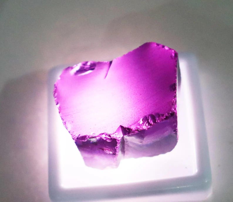 Puede incluir: Una vibrante gema p&uacute;rpura transl&uacute;cida de forma irregular. La gema se exhibe sobre una base cuadrada blanca iluminada, mostrando sus bordes irregulares y la refracci&oacute;n interna de la luz. El color var&iacute;a del violeta intenso a tonos m&aacute;s claros.