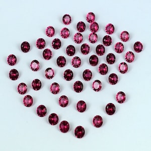 Puede incluir: Una colección de 40 piedras preciosas redondas, facetadas y rosadas. Las piedras preciosas están esparcidas sobre una superficie blanca.