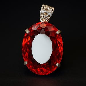 May include: A large, oval-cut red gemstone pendant with a gold-colored bail. The gemstone is faceted, catching the light and creating a brilliant display. The pendant is set against a black background, highlighting its vibrant color and intricate design.