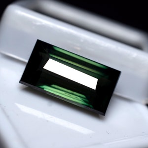 May include: A dark green gemstone with a rectangular cut and a polished finish. The gemstone is set in a white setting.