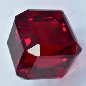 May include: A deep red gemstone with a square shape and many facets. The gemstone is cut in a unique style with a triangular facet in the center.