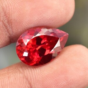 Peut inclure: Une pierre précieuse rouge vif en forme de poire, taillée à facettes pour maximiser la brillance. La gemme est tenue entre les doigts, révélant sa taille et sa forme. Les surfaces taillées reflètent la lumière.