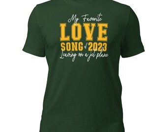 May include: Green t-shirt with the text "My Favorite LOVE SONG of 2023 Leaving on a jet plane" in white and yellow lettering.