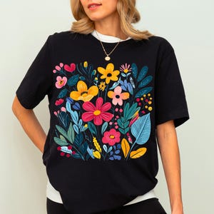 May include: Black t-shirt with a colourful floral print featuring yellow, red, pink, and blue flowers. The flowers are arranged in a bouquet-like design.