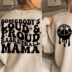 Pu&ograve; includere: Una felpa beige con testo nero che recita "Somebody's Loud & Proud Basketball Mama" e una palla da basket nera con occhi e un sorriso che cola sul retro.
