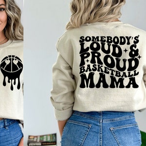 Pu&ograve; includere: Una felpa beige con testo nero che recita "Somebody's Loud & Proud Basketball Mama" in un font retr&ograve;. La felpa &egrave; indossata da una persona con i capelli biondi e jeans blu.