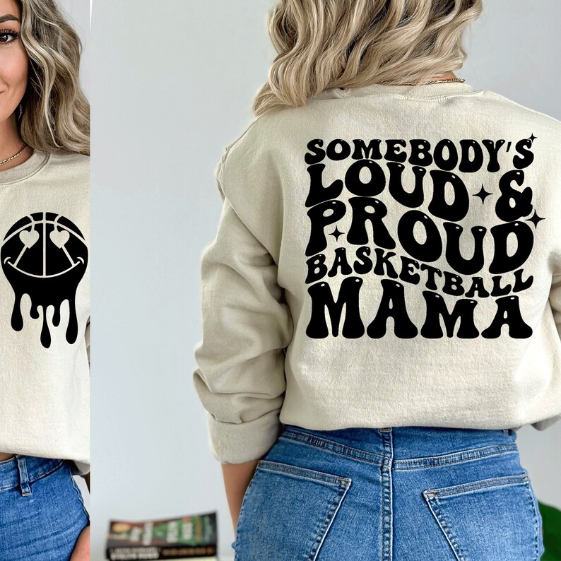 Pu&ograve; includere: Una felpa beige con testo nero che recita "Somebody's Loud & Proud Basketball Mama" in un font retr&ograve;. La felpa &egrave; indossata da una persona con i capelli biondi e jeans blu.