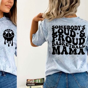 Pu&ograve; includere: Una maglietta blu chiaro con un motivo tie-dye. La parte posteriore della maglietta ha un testo nero che recita "Somebody's Loud & Proud Basketball Mama" in un font retr&ograve;.