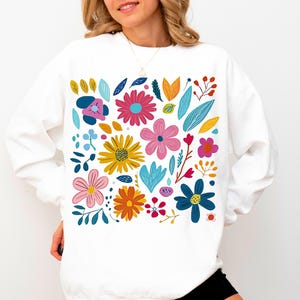 May include: White sweatshirt with a colourful floral print featuring various flowers, leaves, and stems in shades of pink, blue, yellow, orange, and green.