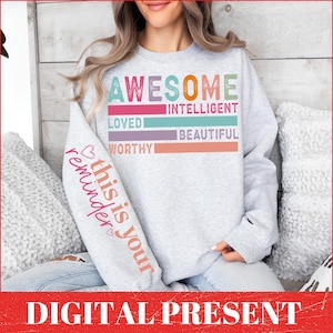 Puede incluir: Sudadera gris con un gráfico colorido que dice "Awesome" y enumera palabras positivas como "Intelligent", "Loved", "Beautiful" y "Worthy". El texto "This is your reminder" está escrito en la manga.