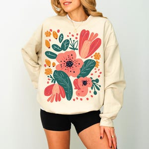 May include: A beige sweatshirt with a floral print featuring pink, red, and green flowers. The flowers are stylised and have a whimsical feel.