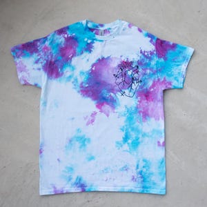 Könnte beinhalten: Weißes T-Shirt mit einem Batikmuster in Blau- und Lilatönen. Das Shirt zeigt eine schwarze Strichzeichnung eines menschlichen Herzens mit Pfeilen. Das T-Shirt ist aus einem weichen Material gefertigt.
