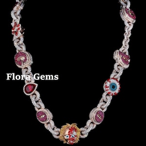 May include: A diamond-encrusted necklace with a chain design. The necklace features red and pink gemstone accents, including a doll face pendant and eye-shaped charms. The text "Flora Gems" is visible.