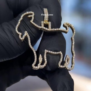 May include: A gold-toned pendant in the shape of a goat outline, encrusted with small clear stones along the entire perimeter. The pendant features a bail at the top and includes the text Flora Gems printed in small black letters near the center.