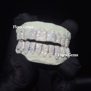 以下が含まれることがあります： ダイヤモンドのパターンが施されたホワイトゴールドのグリルで、右下に「Flora Gems」の文字があります。