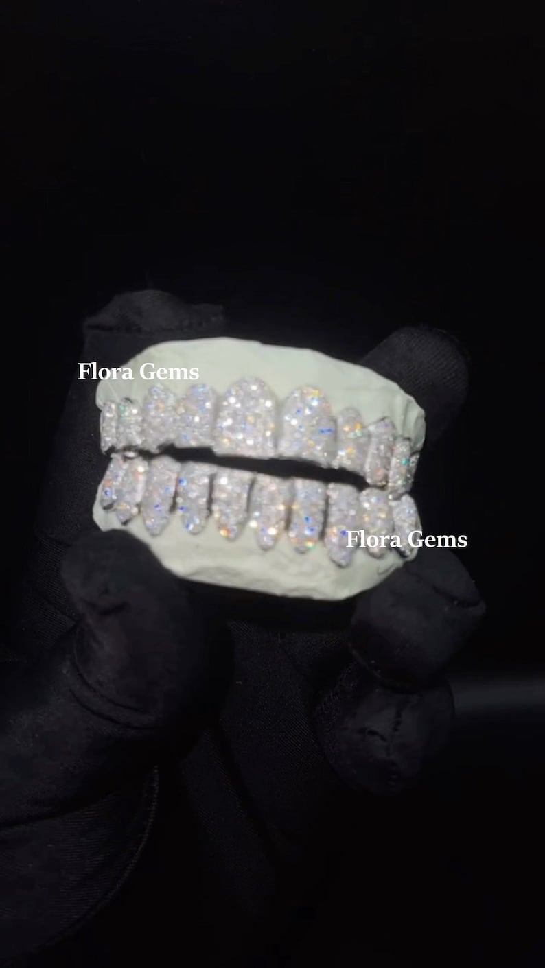 以下が含まれることがあります： ダイヤモンドのパターンが施されたホワイトゴールドのグリルで、右下に「Flora Gems」の文字があります。