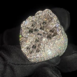 May include: A silver-toned ring featuring a large, cross-shaped centerpiece densely encrusted with numerous small, sparkling clear stones. The surface of the ring is fully paved with these stones, and the text Flora Gems is visible in the center.