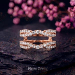 May include: A rose gold ring guard featuring two curved bands connected by vertical bars. Each band is encrusted with a row of small, round, clear stones in a pavé setting. The design creates a symmetrical, wave-like appearance with a central gap.