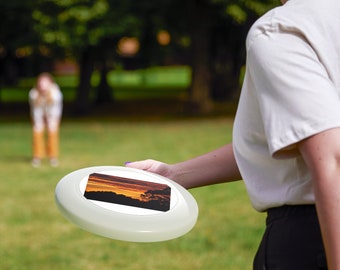 Può includere: Un frisbee di plastica bianca con una scena di tramonto colorata stampata sulla parte superiore. Il frisbee è tenuto da una mano.