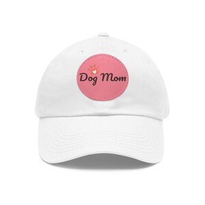 Pode incluir: Boné de beisebol branco com um patch circular rosa que diz "Dog Mom" com um design de pata e coração.
