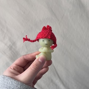 Puede incluir: Una pequeña figura verde de una persona que lleva un gorro rojo de punto con un pompón. La figura está sentada en una postura meditativa con las manos juntas frente al pecho.